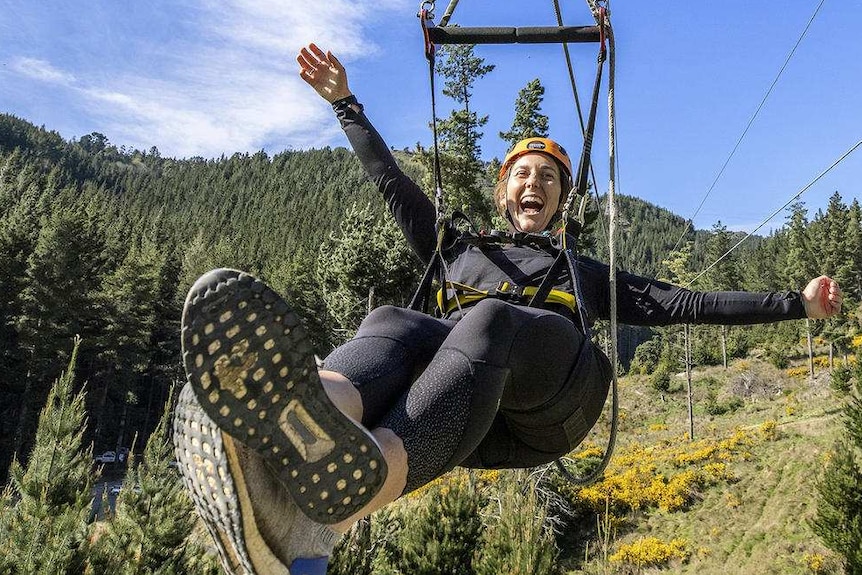 A smiling woman holds her hands out as she ziplines over a green forest.