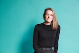 Portrait of choreographer Stephanie Lake, a white woman sitting on a stool in black, teal backdrop, looking serious