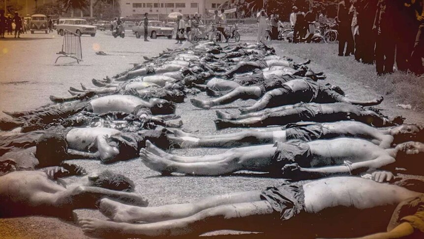 A black and white photo shows a street scene showing two rows of dead bodies as a crowd watches on.
