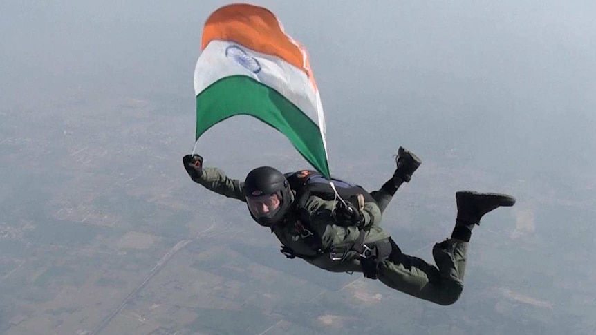 A man in a green flight suit and helmet holds an Indian flag above his head as he skidives