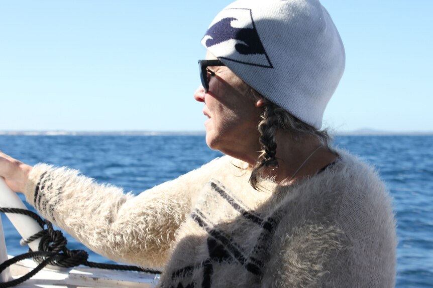 A woman wearing a beanie, jumper and sunnies sits on a boat looking out to the ocean.
