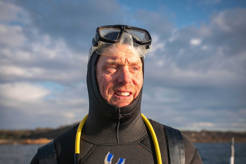 A man in a diving wetsuit, with goggles on top of head and oxygen tube around neck, stares into the distance with morning light.