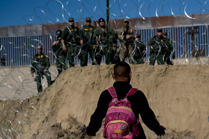 A man wearing a backpack holds his hands out to show they are empty as nine armed US border guards look on through razor wire.