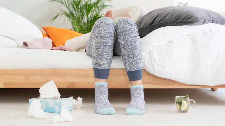 Legs clad in tracksuit pants and fluffy socks can be seen next to tissues and tea as someone lies back on a bed.