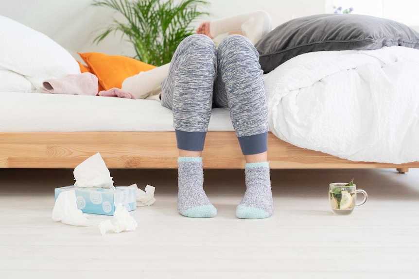 Legs clad in tracksuit pants and fluffy socks can be seen next to tissues and tea as someone lies back on a bed.