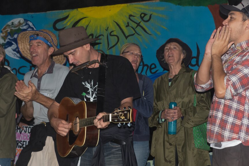 A man play guitar and a harmonica surrounded by people cheering.