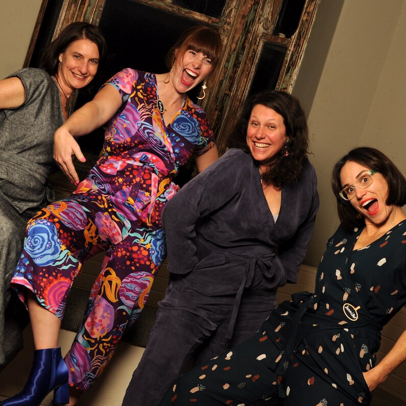 Four women wearing jumpsuits in identical designs but different fabrics smile at the camera.