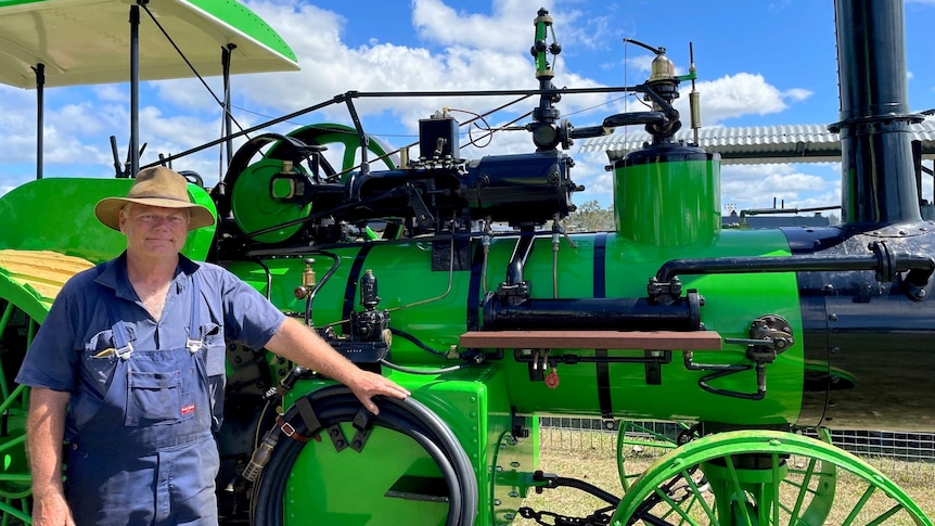 A man wearing overalls and a hat stands next to a bright green steam powered tractor.