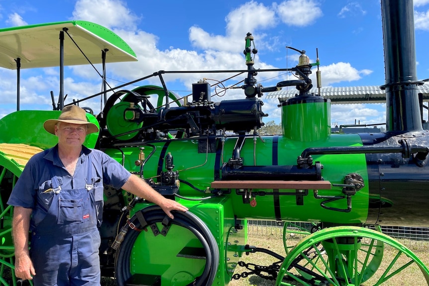 A man wearing overalls and a hat stands next to a bright green steam powered tractor.