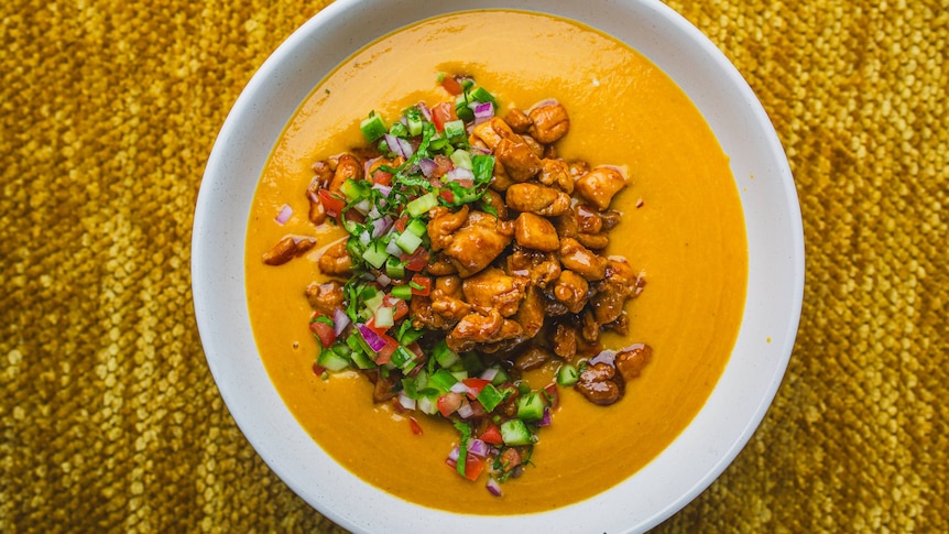A bowl of creamy red lentil soup on a table, topped with sticky chicken, chopped tomatoes , cucumber and herbs.