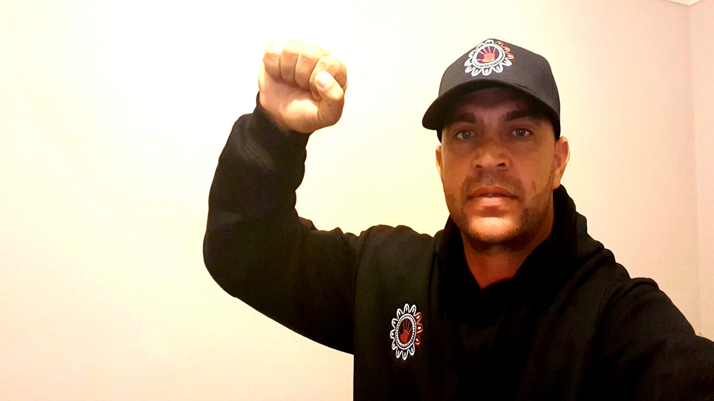 Jeffery Amatto holding up his fist, wearing a black cap and shirt with logo on it