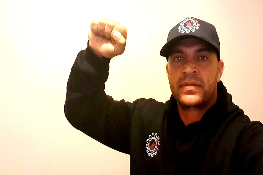 Jeffery Amatto holds his fist up, wears a black cap and shirt with logo on it