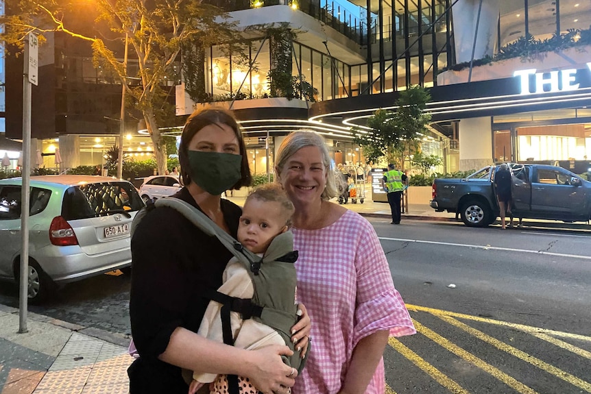 A woman wearing a black shirt and mask has a baby strapped to her chest standing next to a woman in a pink gingham shirt.