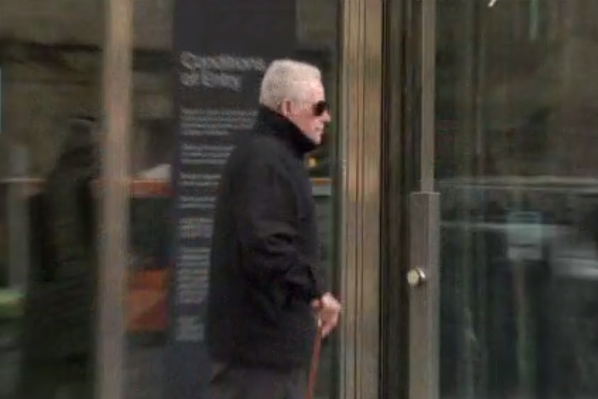 An elderly man dressed in black walks into court with the aid of a walking stick.