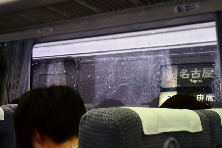 Inside of a train, the back of two people's heads, a sign in Japanese and English for 'Nagoya'