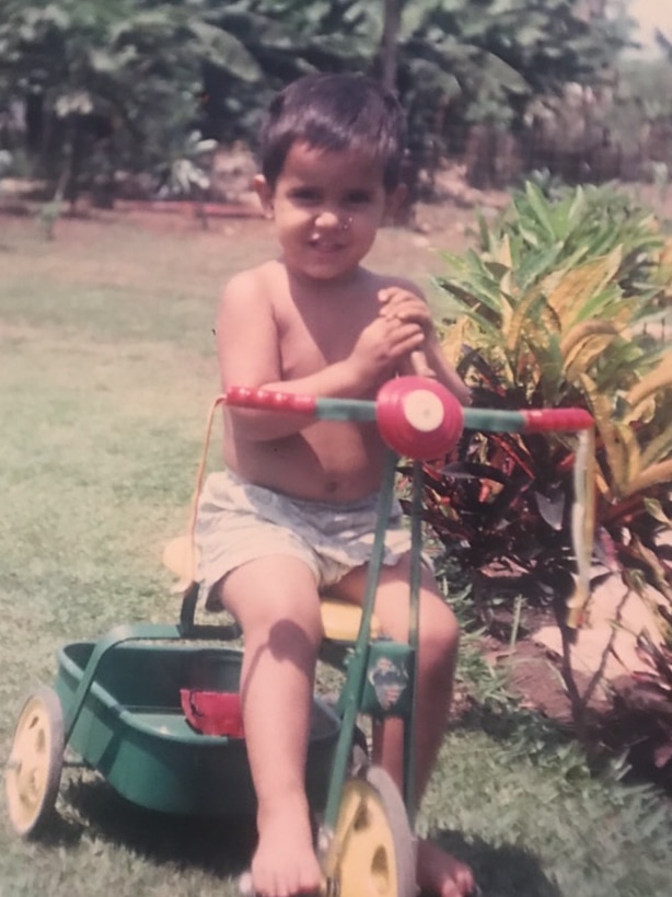 An old photo of a young boy in his backyard, sitting on a bike and smiling.