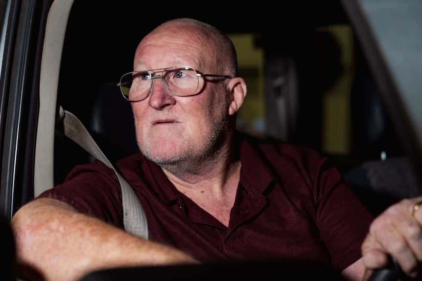 a bald man with glasses and a maroon shirt looks over his shoulder as he sits at the front wheel of his vehicle