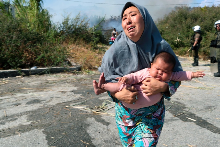 A migrant woman wearing a headscarf holds her baby as she runs to avoid a fire in Lesbos.