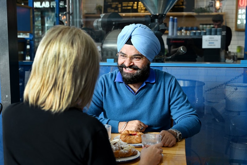 A man in a blue turban seated in a cafe at a table with a woman whose face is not shown.