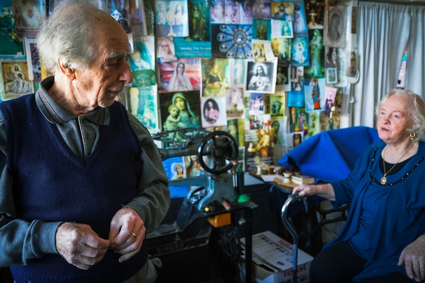 An elderly couple look at each other with posters on the wall in the background