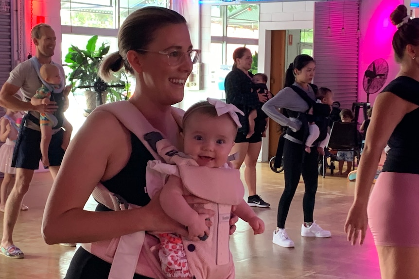 Young mum in exercise gear smiling and dancing with daughter in a pouch