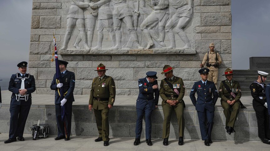 A group of men stand in green and blue uniforms in front of a stone relief depicting soldiers.