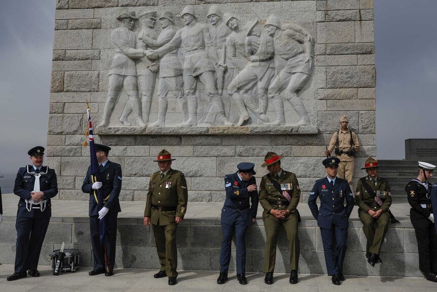 A group of men stand in green and blue uniforms in front of a stone relief depicting soldiers.