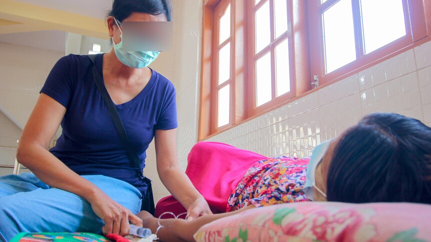 A woman in a face mask and blue t-shirt treats a woman lying on a bed 