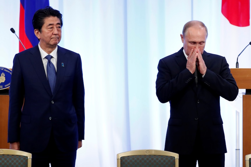 블라디미르 푸틴이 명상을 하는 동안 얼굴에 손을 얹고 아베 신조가 멀리서 바라보고 있다.