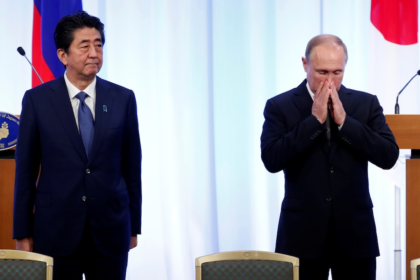 Vladimir Putin putting his hands on face in contemplation as Shinzo Abe stares off in the distance