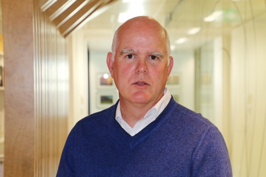 A close-up shot of a man wearing a navy blue v-neck jumper and white shirt.