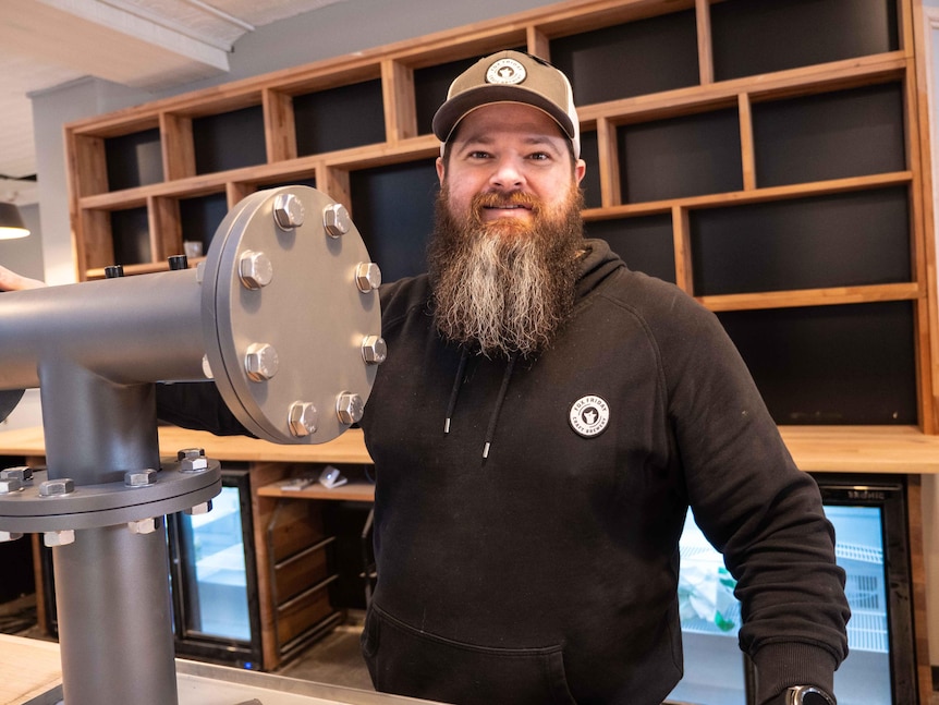 A man with a beard wearing a cap standing behind a bar looking at the camera