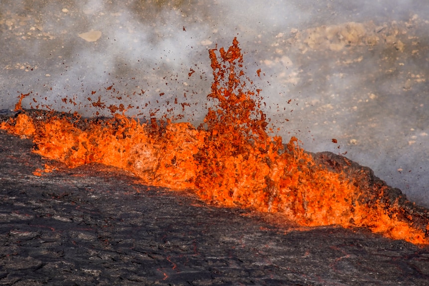 Dark red lava explodes from black rock in a straight line downhill as smoke rises from it 