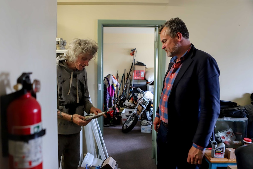 One man wearing dark suit jacket and checkerd shirt stands opposite grey haired man with grey jumper standing in apartment room