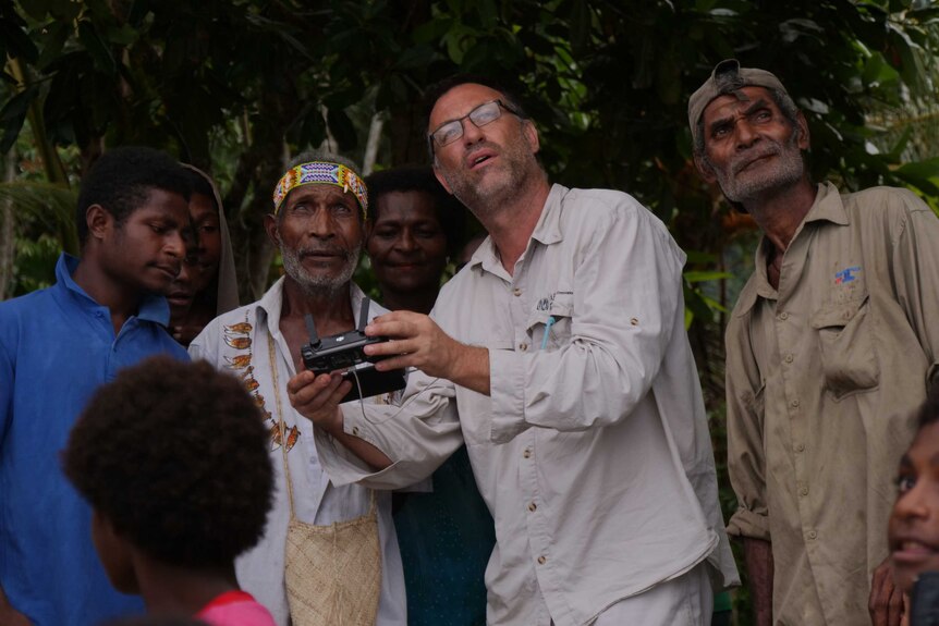 Castellaro holding drone controls and showing them to a group of PNG locals who look at screen or up at sky.