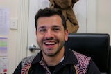 A man with dark hair and facial hair smiles at the camera, sitting at his desk. 