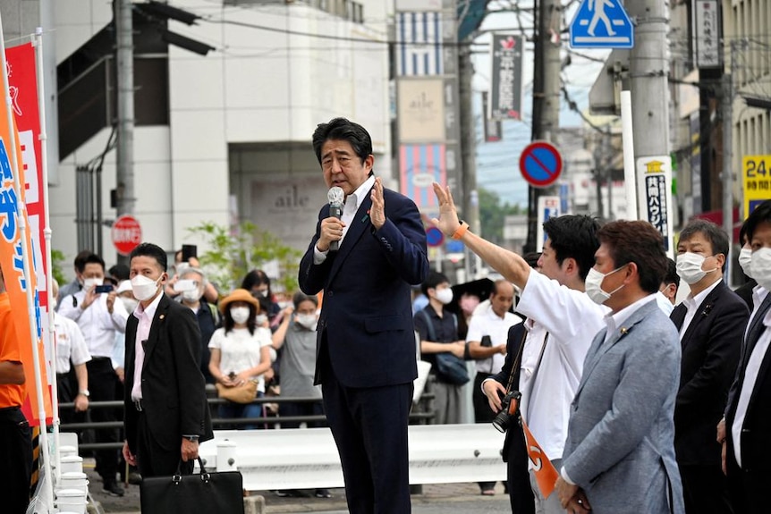 A japanese politician in a suit holding a microphone stands on a platform on a city street with people in masks surrounding him 