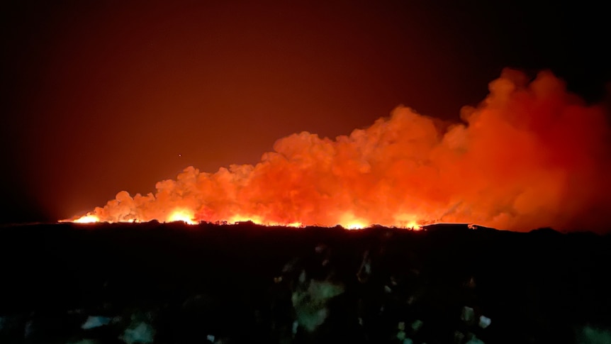 A bushfire burns on the horizon at night, lighting up the sky orange.