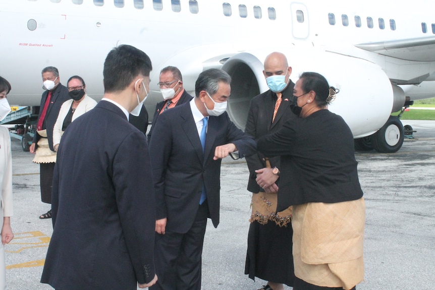 Chinese Foreign Minister wears mask and does an elbow bump in greeting a woman from Tonga with airplane in the background.