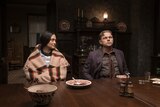 A film still showing a woman wearing a Native American shawl sitting at a dining table next to a white man in a jacket and tie