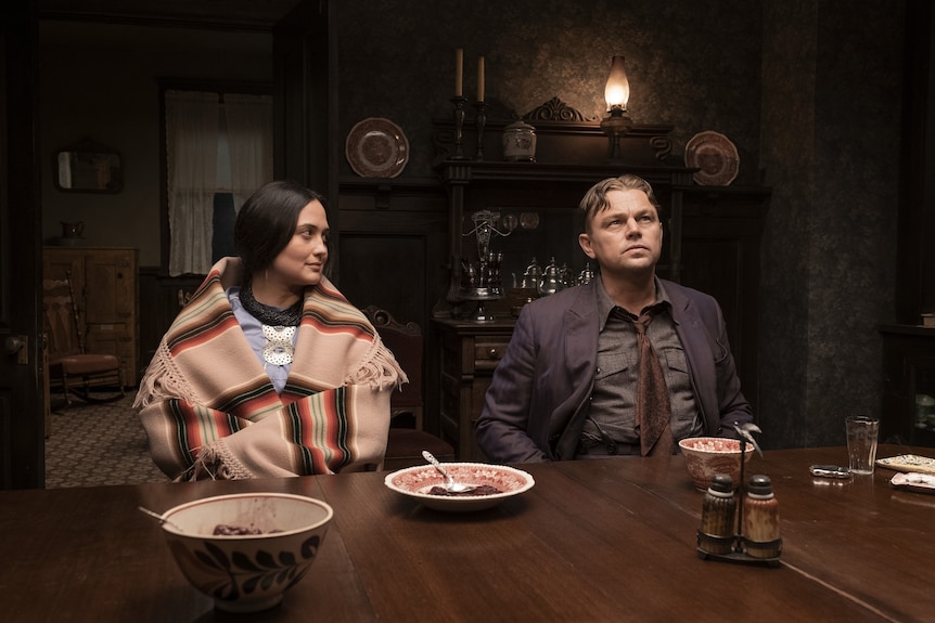 A film still showing a woman wearing a Native American shawl sitting at a dining table next to a white man in a jacket and tie