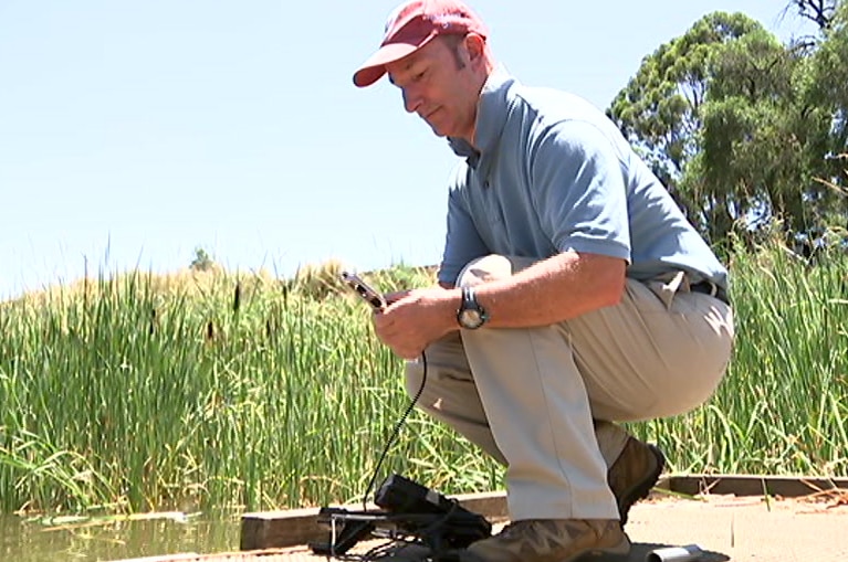 A man holds a device in his hands while kneeling on a riverbank