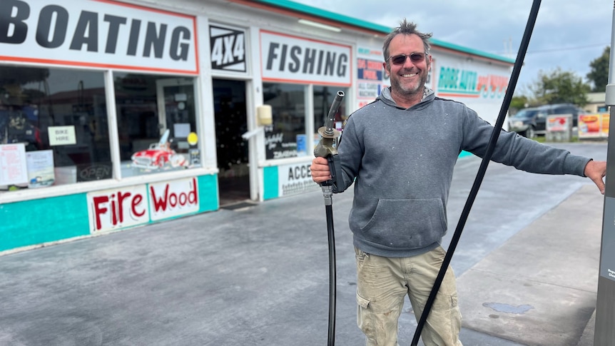 A smiling man in a grey jumper stands next to a petrol pump 