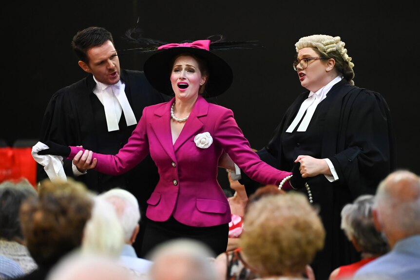 A woman wearing a pink jacket and hat sings with her arms spread while two other actors dressed as barristers look on