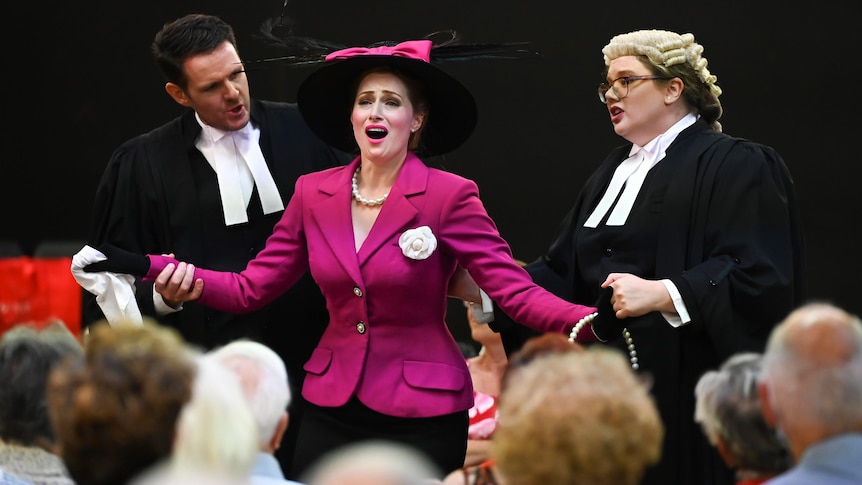 A woman wearing a pink jacket and hat sings with her arms spread while two other actors dressed as barristers look on
