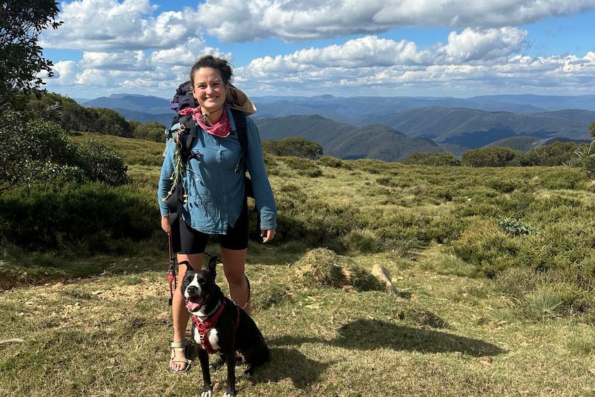 A woman in hiking gear smiling with a dog at her feet, with a mountain range in the background. 