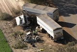 The wreckages of a car and a semi-trailer lie on the side of a country road as two police officers examine the scene.