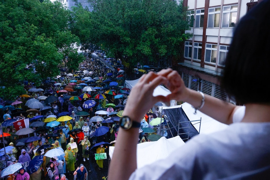 A woman holds up a love heard with her hands in front of a crowd of protesters holding umbrellas.