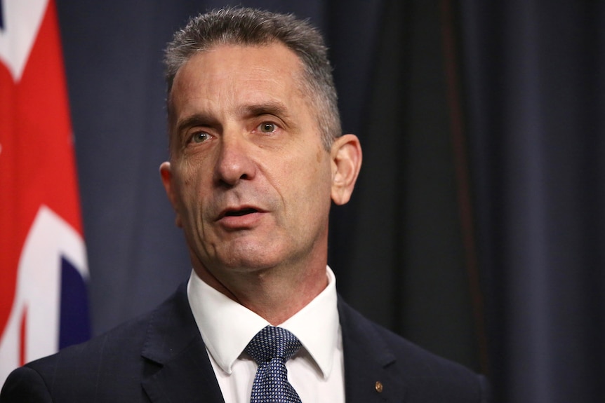 A close-up shot of a man in a suit and tie speaking at a media conference indoors in front of an Australian flag.