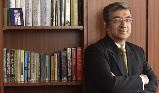 Bespectacled man wearing suit and tie standing with arms folded in front of book chest